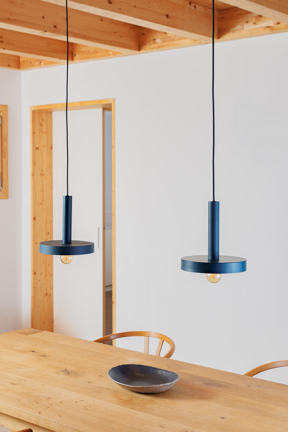Two blue pendant lights hanging above a wooden dining table with a white wall and wooden beams in the background.