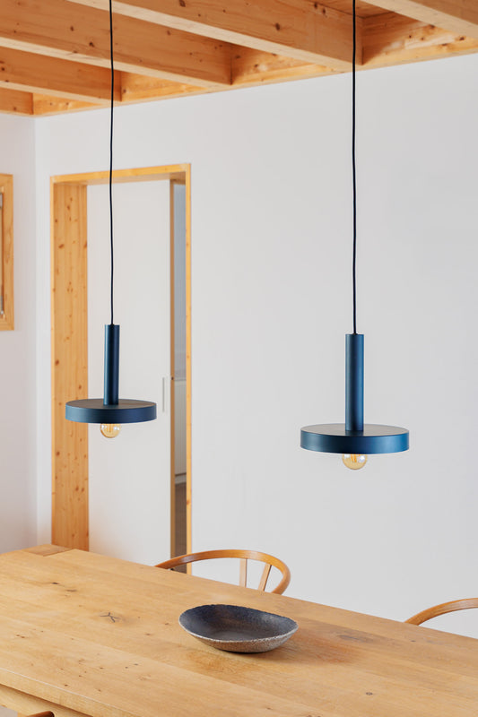 Two blue pendant lights hanging above a wooden dining table with a white wall and wooden beams in the background.