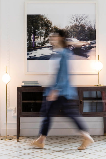 Person walking past a television displaying a snowy scene with a car, in a room with a wooden cabinet and floor lamp.