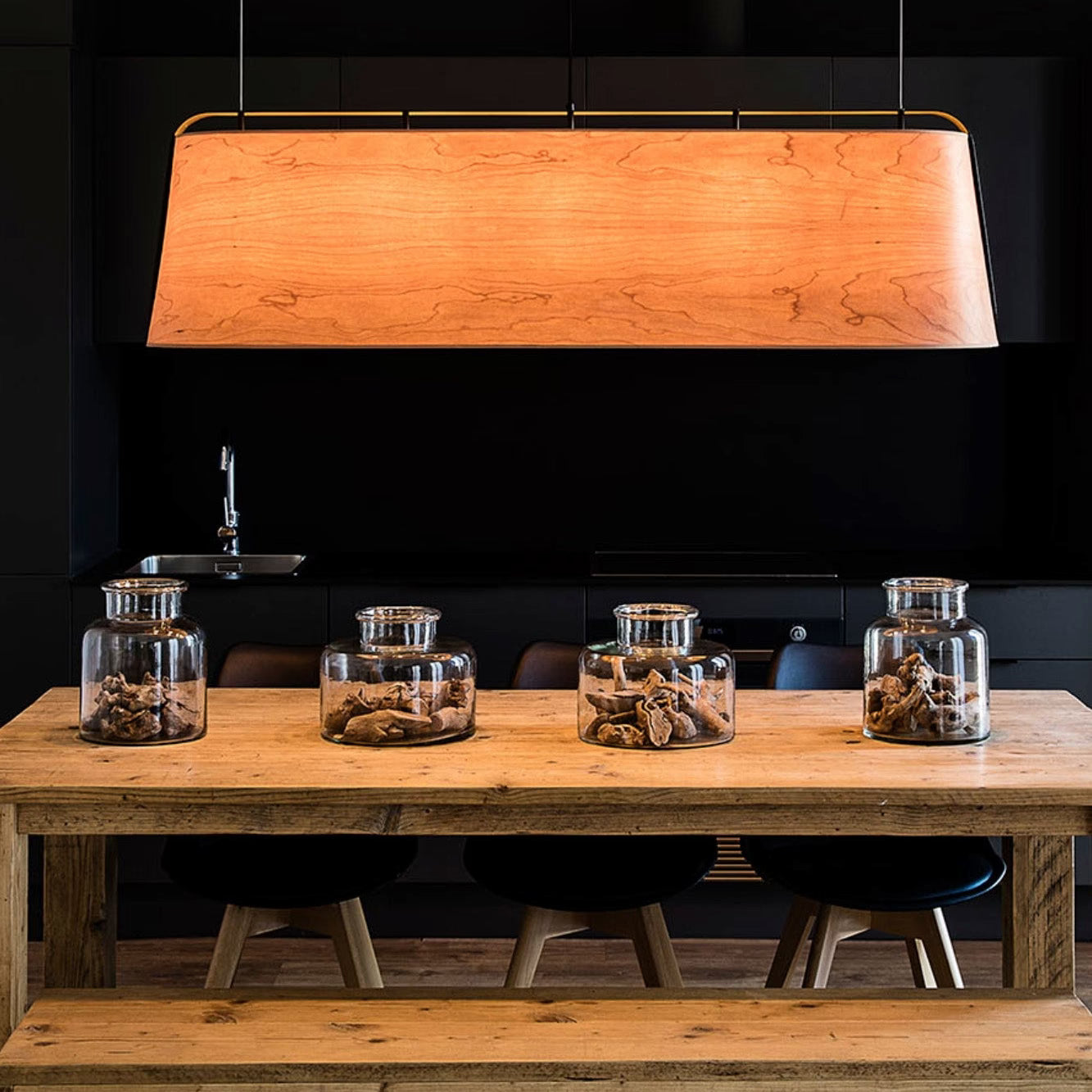 A rectangular wooden pendant light with a warm glowing wood-grain finish hangs over a rustic wooden dining table in a dark modern kitchen setting. Three clear glass jars and black stools are visible on the table.