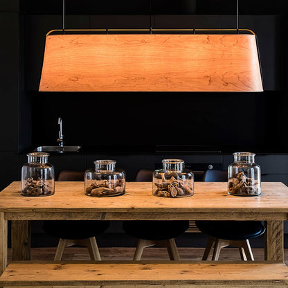 A rectangular wooden pendant light with a warm glowing wood-grain finish hangs over a rustic wooden dining table in a dark modern kitchen setting. Three clear glass jars and black stools are visible on the table.