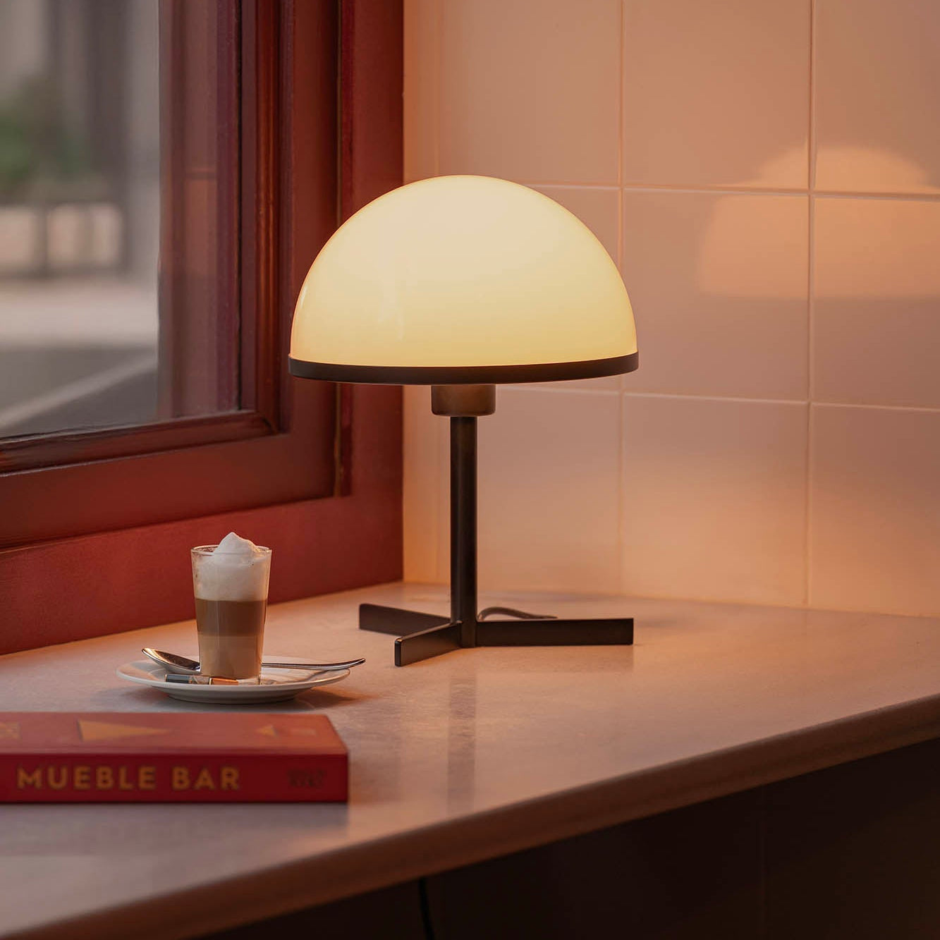 Table lamp on a surface next to a book and a small plate with a dessert near a window.