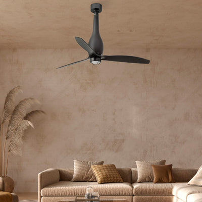 A matte black modern ceiling fan with three blades hanging in a minimalist living room with textured beige walls, a beige sectional sofa, and a low wooden coffee table.