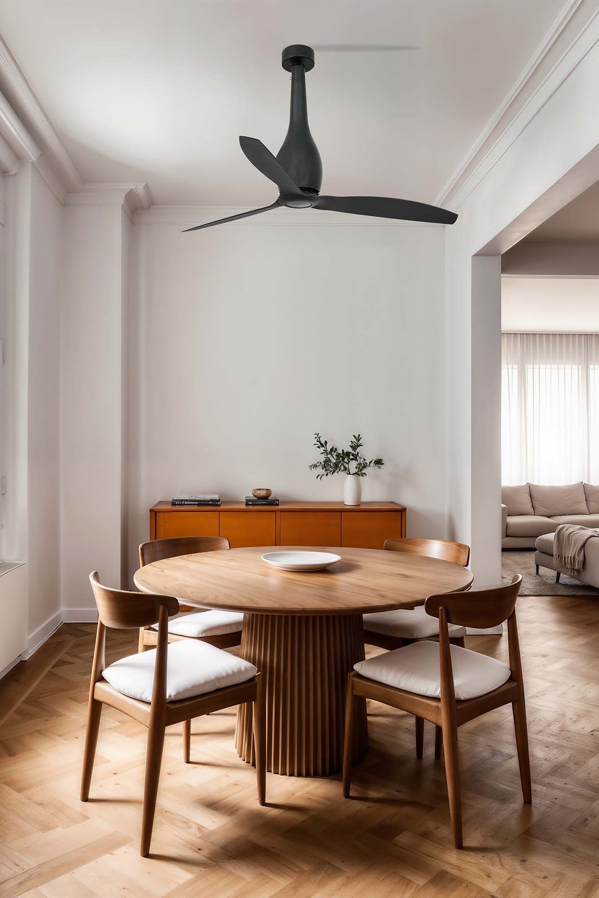 A modern black ceiling fan with a sculptural, multi-leaf design hangs in a bright, minimalist dining room with a round wooden table and four matching chairs.