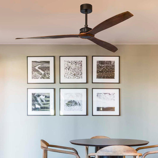A modern dining room scene featuring a dark wood ceiling fan mounted on a light beige wall above a round black table and wooden chairs. Six framed black-and-white artworks are arranged on the wall.