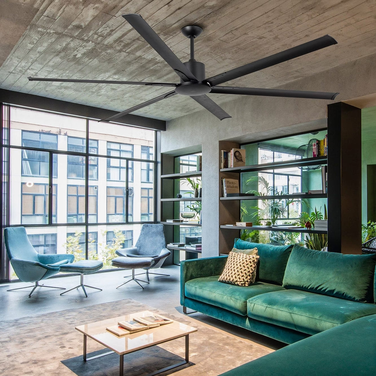 A modern living room interior featuring a dark teal velvet sofa, two matching teal lounge chairs, a light wood coffee table, and a large ceiling fan mounted on a concrete ceiling. Large windows look out onto a city building, and shelving with plants is visible along the right wall.