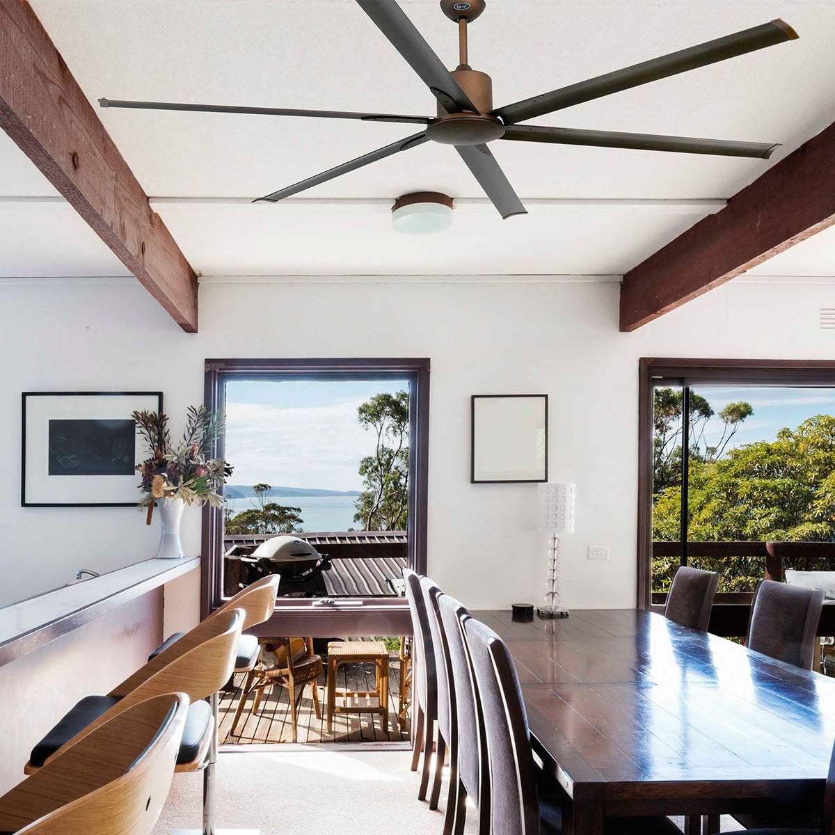 A modern dining room interior with a dark wood dining table and chairs, a bar counter on the left, exposed wooden ceiling beams, and large windows overlooking a tropical outdoor scene with the ocean and trees.