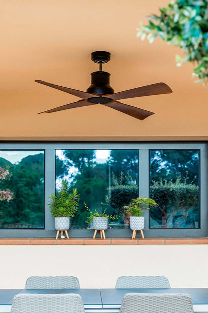 A black ceiling fan with dark brown blades installed on a terracotta-colored ceiling, shown in a modern patio or outdoor dining setting with large windows, potted plants, and woven chairs.