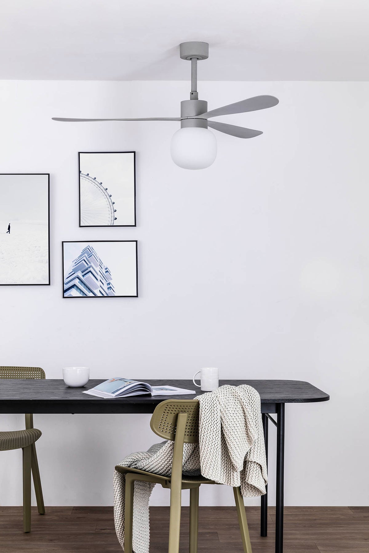 A modern gray ceiling fan with a frosted white globe light hangs above a dining table in a minimalist room with framed wall art and chairs.