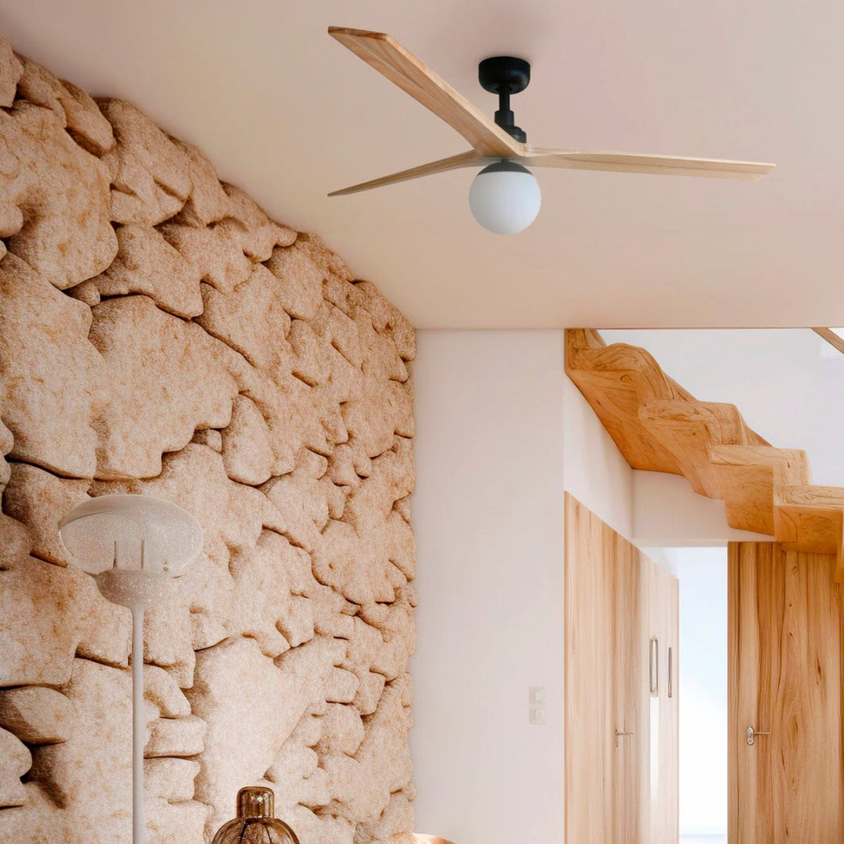A modern ceiling fan with light wood blades and a white globe light fixture, installed in a minimalist interior with a textured stone wall and wooden staircase.