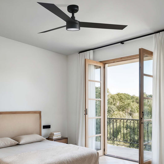 A modern bedroom featuring a light wood bed frame with beige bedding, a black ceiling fan, and open French doors leading to a balcony with a view of green trees.