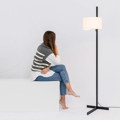 Woman sitting on a white cube with a floor lamp next to her against a light gray background