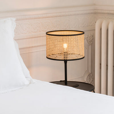 Table lamp with a woven shade on a nightstand next to a bed.