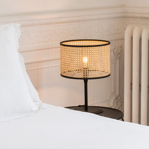 Table lamp with a woven shade on a nightstand next to a bed.