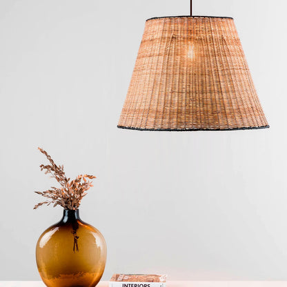 A woven natural fiber pendant lampshade hangs from a black cord against a light gray wall, with a brown glass vase and dried stems on a tabletop below.