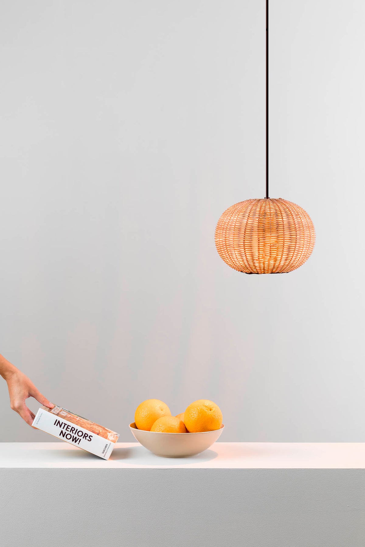 Woven pendant light hanging above a bowl of oranges on a white surface with a gray wall background.