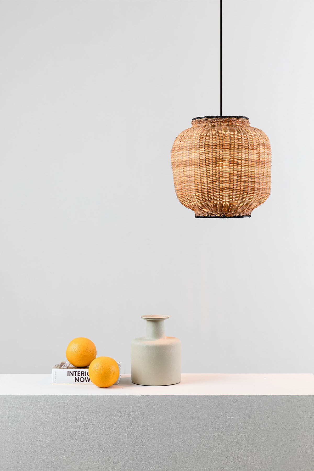 Woven pendant lamp hanging above a white surface with a small ceramic vase and oranges.