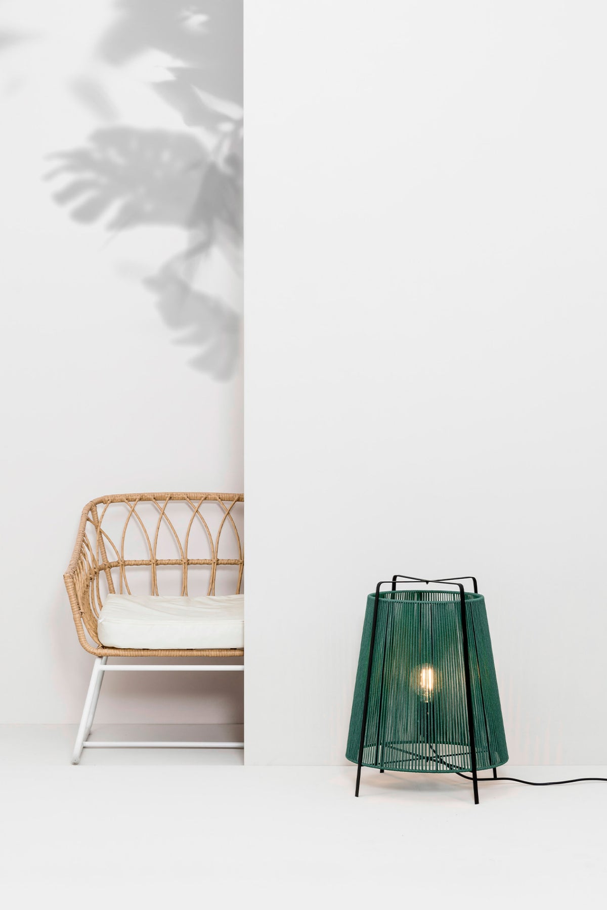 Modern green table lamp with a wicker chair against a white wall.