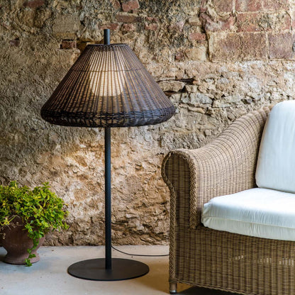 A tall floor lamp with a dark woven conical shade and black metal stand, placed beside a wicker armchair with white cushions against a rustic stone wall.