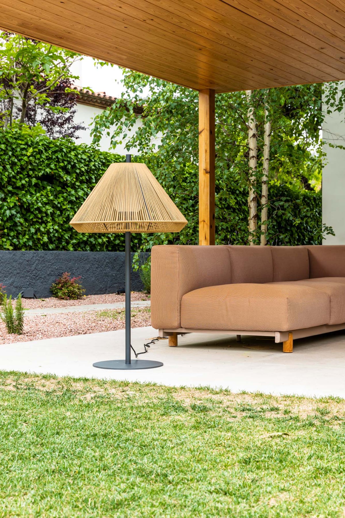 Brown outdoor sofa under a wooden canopy with a lamp in a garden setting.
