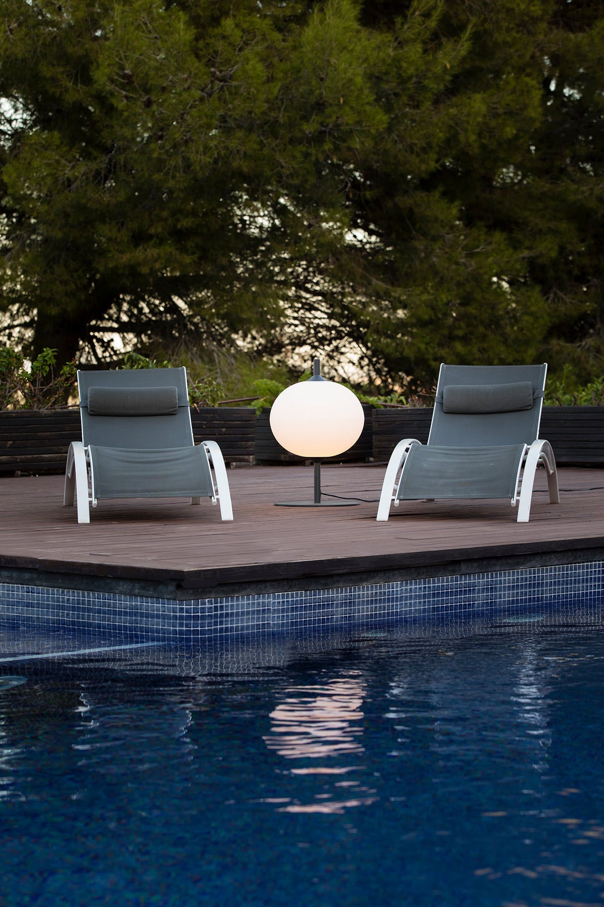 Two lounge chairs by a pool with a spherical lamp between them, surrounded by trees.