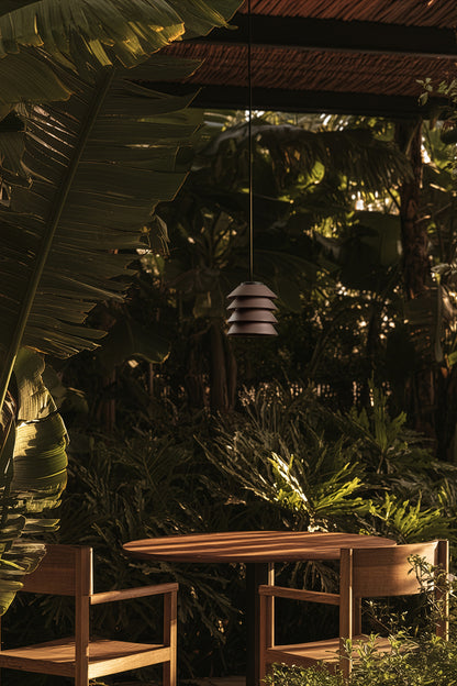 Outdoor setting with a table and chairs under a hanging lamp, surrounded by lush greenery.