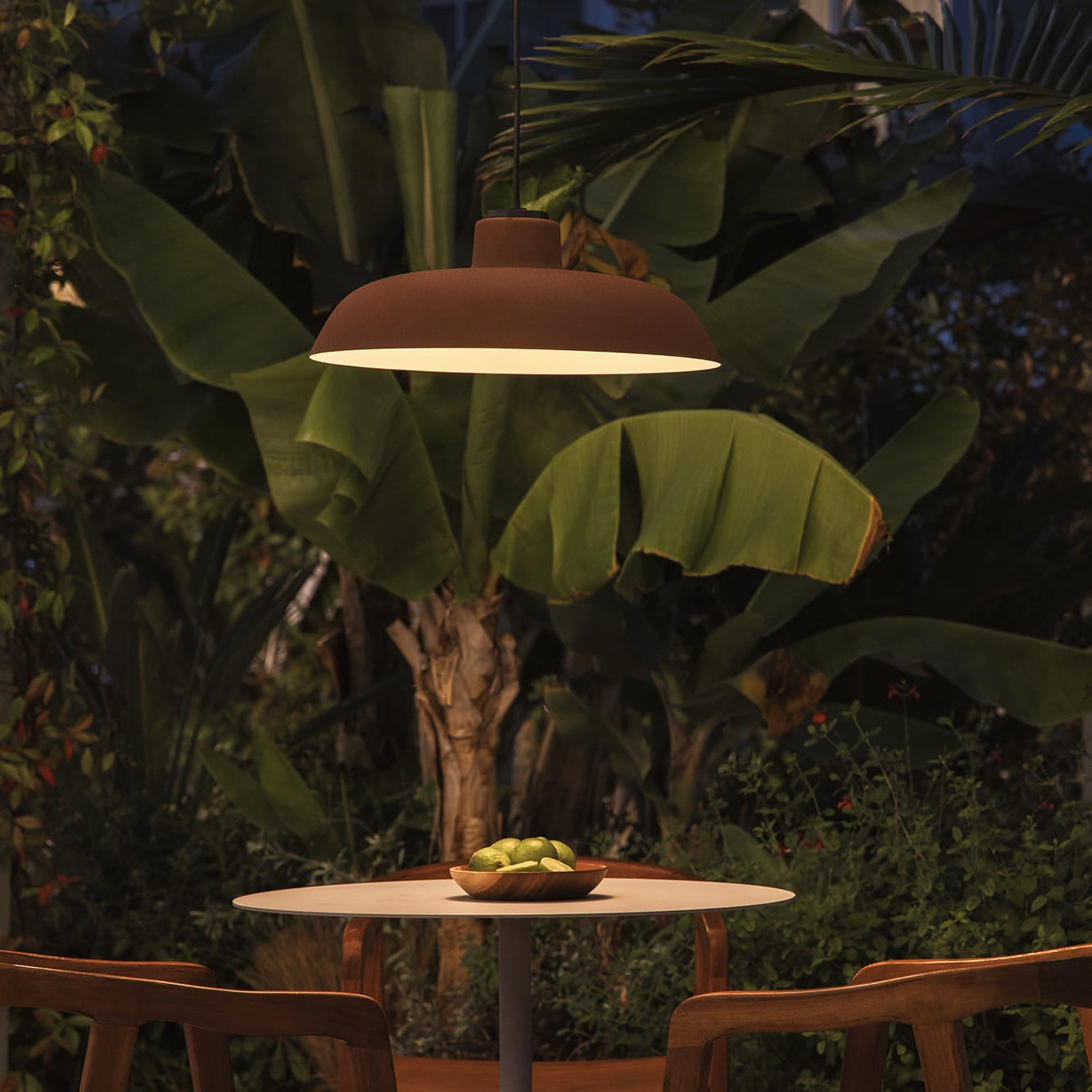 Outdoor setting with a hanging lamp, table, and chairs under a banana leaf at night.