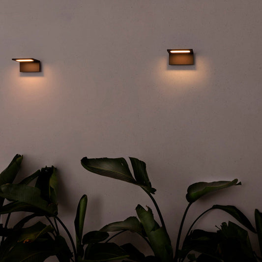 Two outdoor wall lights on a white wall with plants in the foreground.