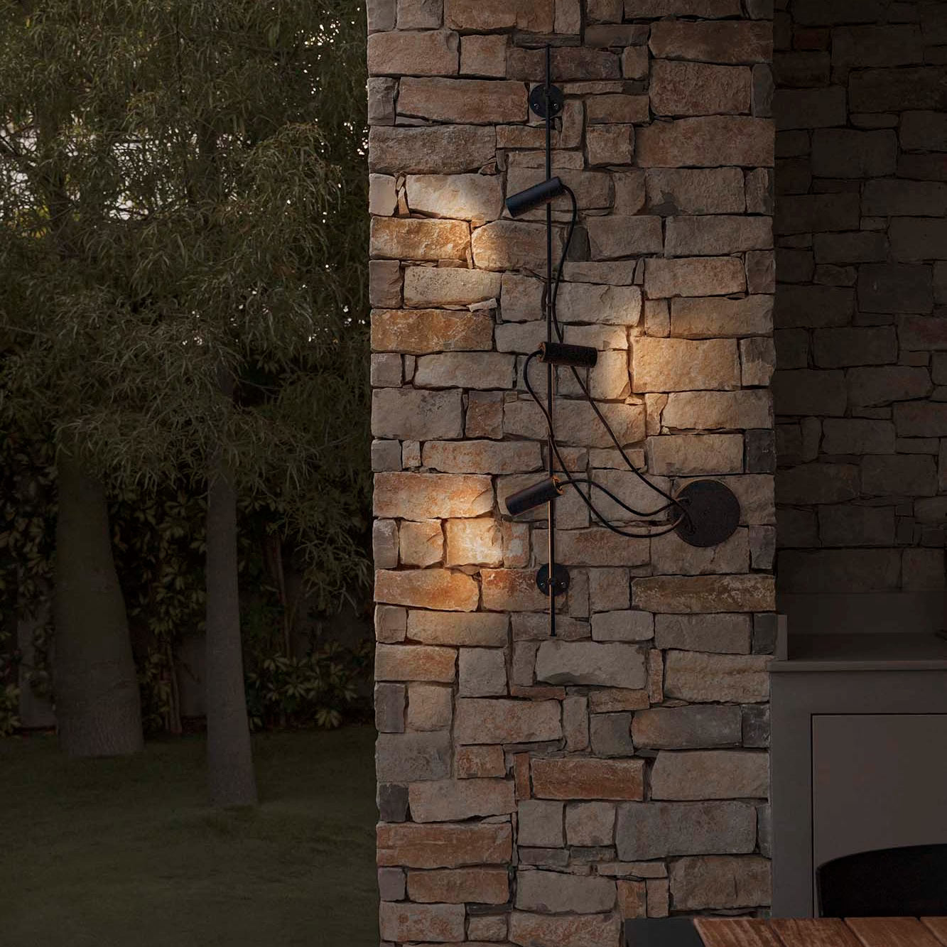 A modern black outdoor wall-mounted light fixture with multiple adjustable LED spotlights and a circular diffuser, shown installed on a stone pillar at night, casting warm light onto the stone surface.