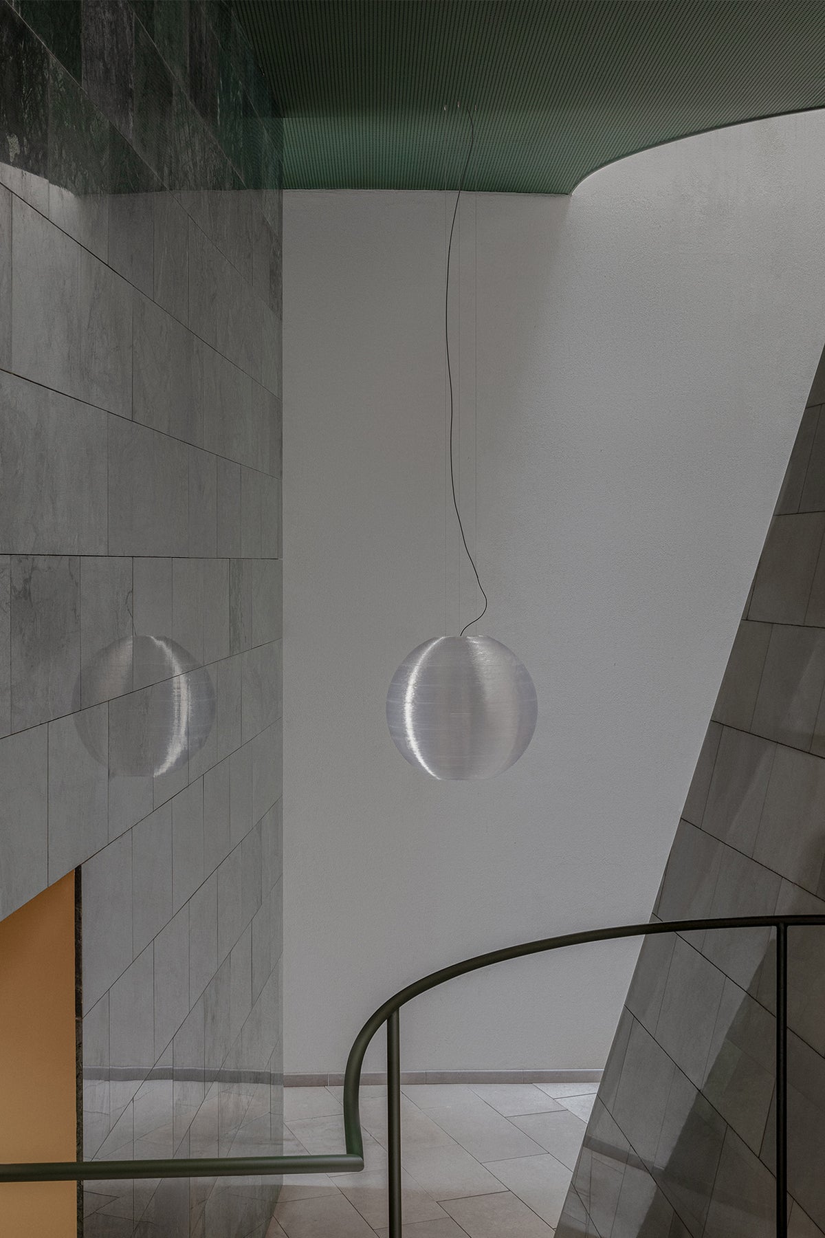 A modern spherical pendant light with a textured, metallic silver finish hangs in a minimalist interior space with gray tiled walls and a curved black railing.