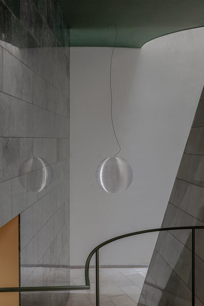 A modern spherical pendant light with a textured, metallic silver finish hangs in a minimalist interior space with gray tiled walls and a curved black railing.