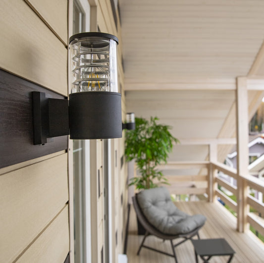 Black outdoor wall light fixture on a house exterior with a chair and plant in the background.