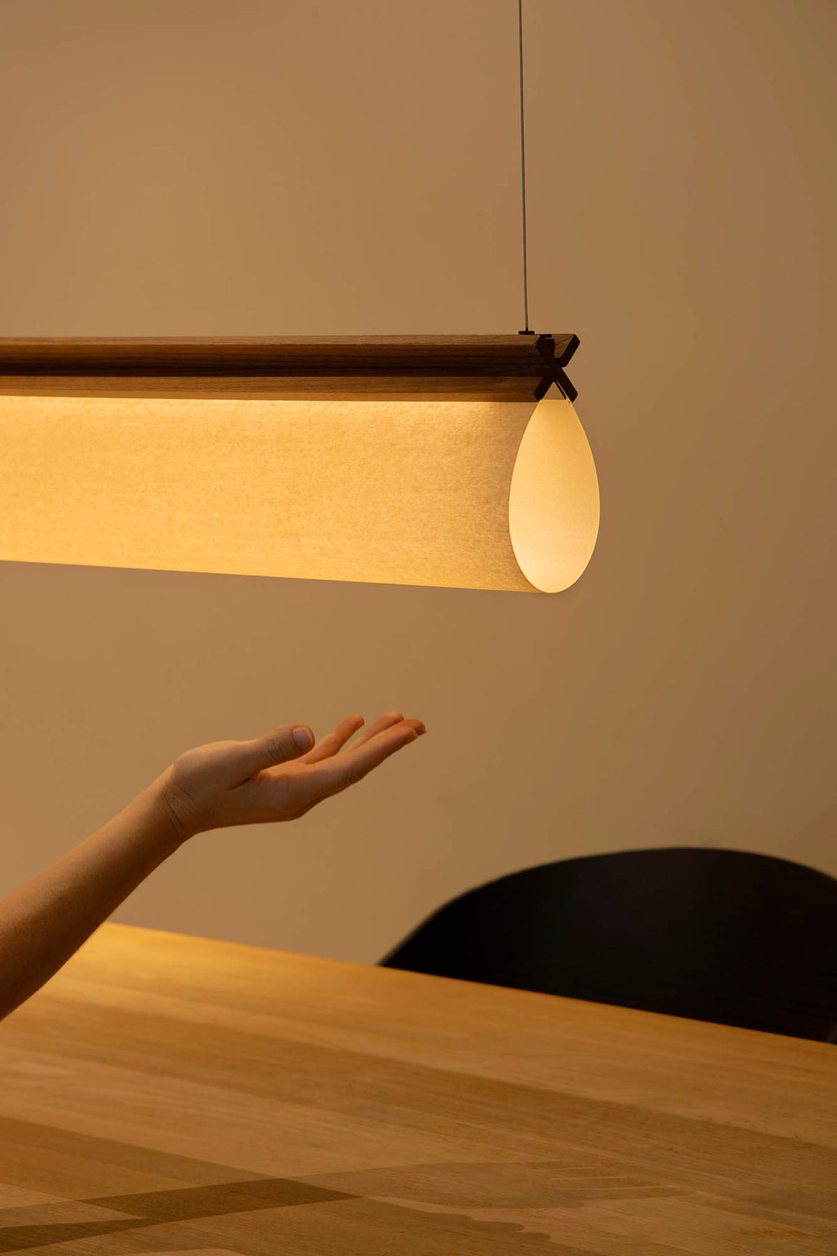 A modern linear pendant light with a warm glowing fabric shade and dark wood trim, shown hanging above a wooden table in a minimalist interior. A person's hand is reaching toward the light, and a dark chair is visible in the background.