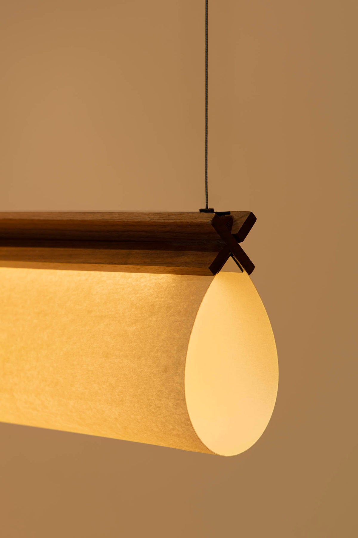 A modern cylindrical pendant light with a warm glowing beige fabric shade, a dark wood top bar, and a black hanging cord against a solid warm beige background.