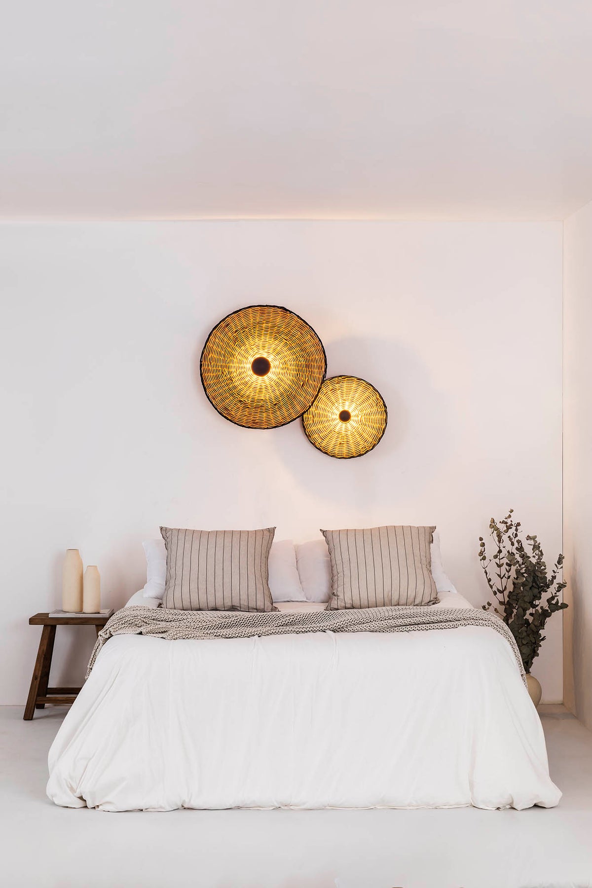 A minimalist bedroom scene with a neatly made bed in white bedding, two striped pillows, a small wooden side table with candles, and two round woven wall lamps mounted on a white wall above the bed.