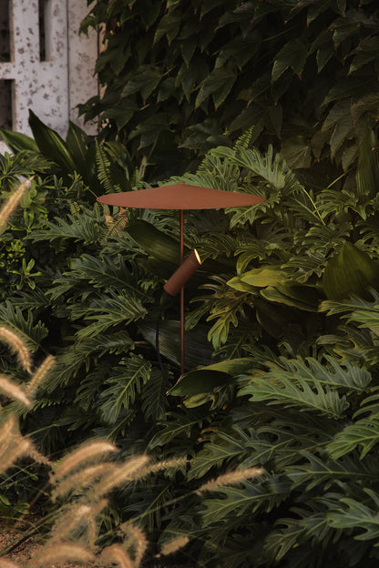 A modern minimalist outdoor table lamp with a terracotta-colored umbrella canopy and a matching cylindrical shade, set against a dense background of lush green foliage.