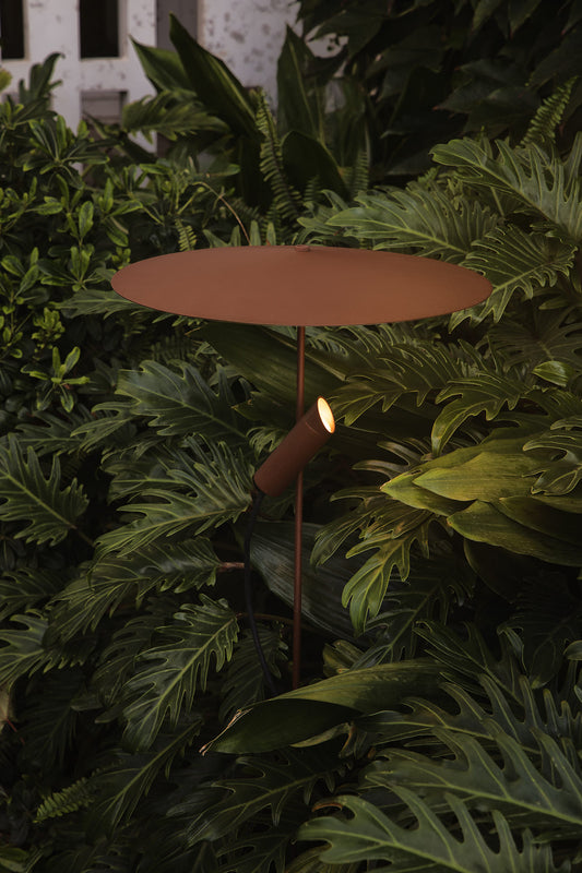 A modern outdoor garden light with a round rusted metal top and a directional spotlight, nestled among lush green tropical foliage.