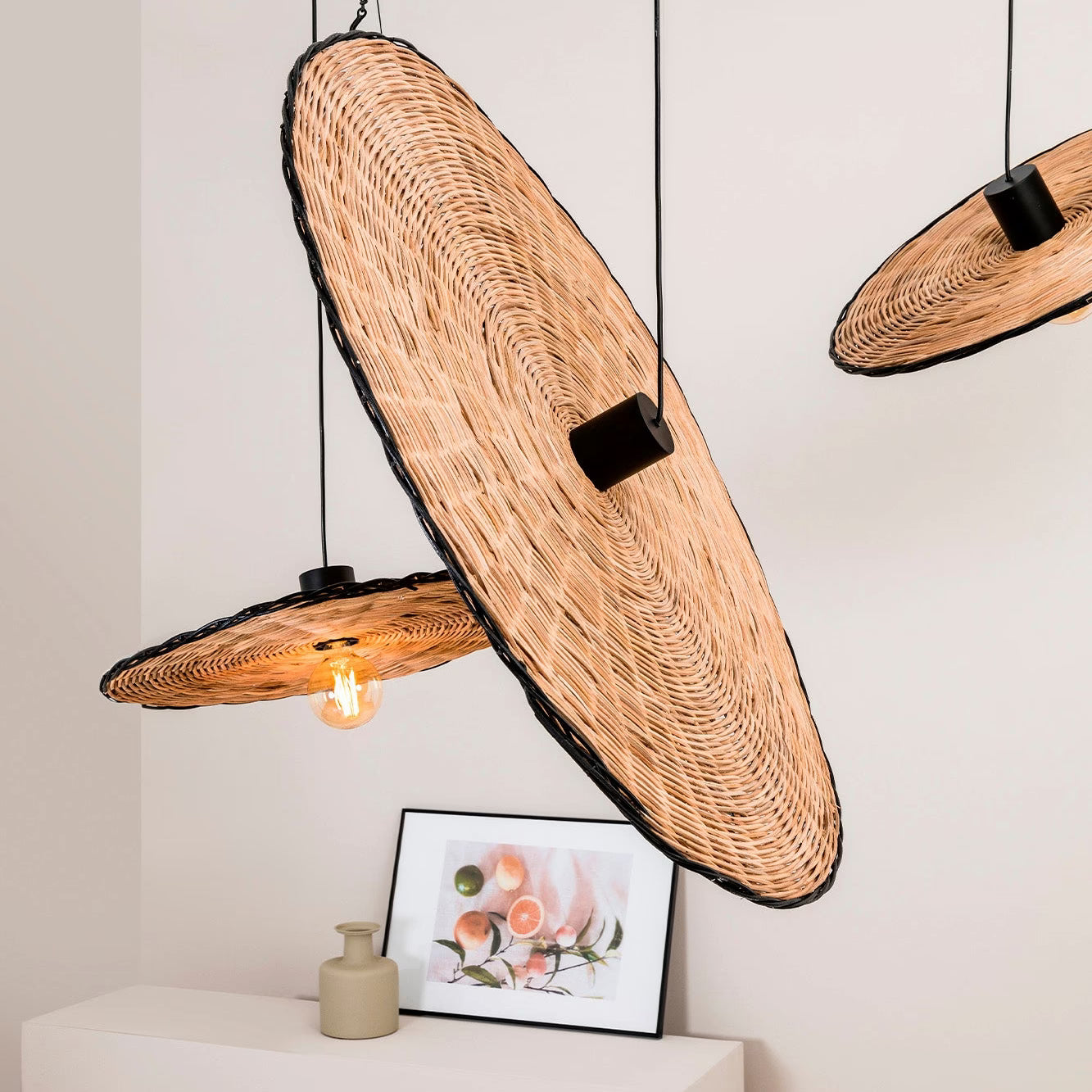 Three woven wicker pendant light shades with black metal hardware and exposed Edison bulbs, hanging in a minimalist room setting above a white console table with a small framed print.