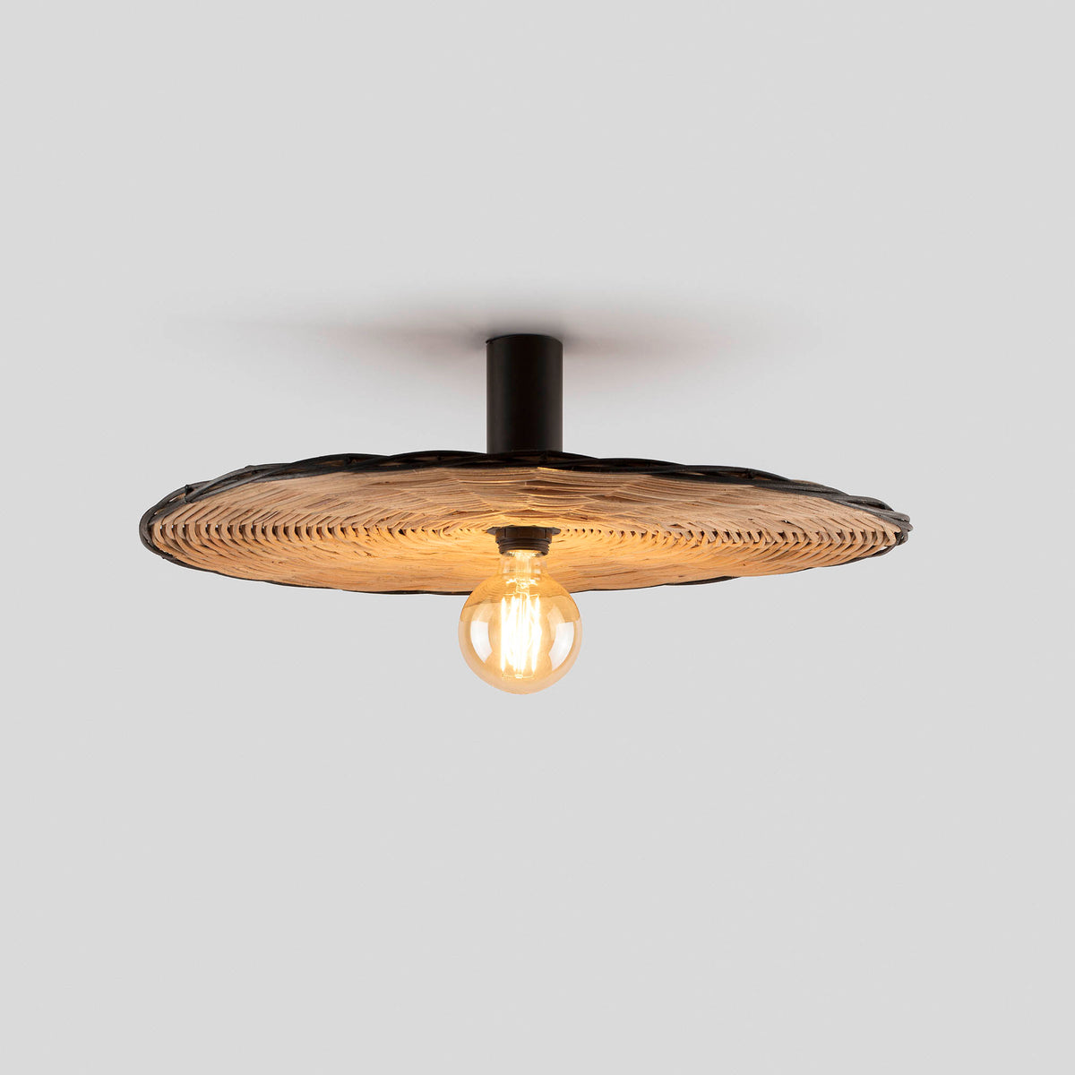 The Faro Barcelona Costas 600 lamp mounted as a flush ceiling light. It features a wide, woven natural rattan shade with a dark edge and a matte black cylindrical base, illuminating the space with an exposed decorative Edison-style bulb.