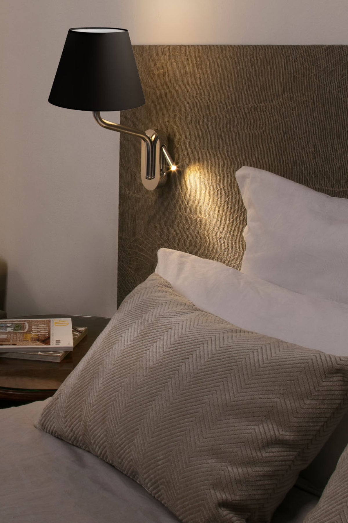 Bedroom scene with a lamp on the wall, pillows, and a magazine on a nightstand.