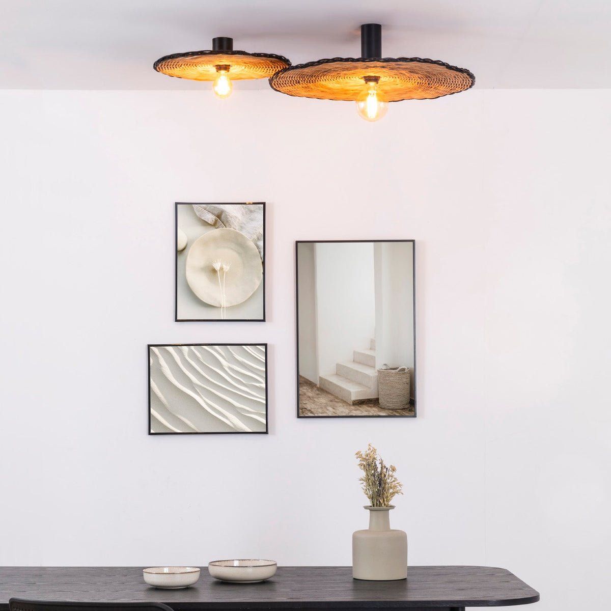A modern dining room featuring two Faro Barcelona Costas rattan flush mount ceiling lights. The woven natural shades cast a warm glow from their exposed bulbs above a minimalist black dining table, complementing a stylish gallery wall of neutral artwork.