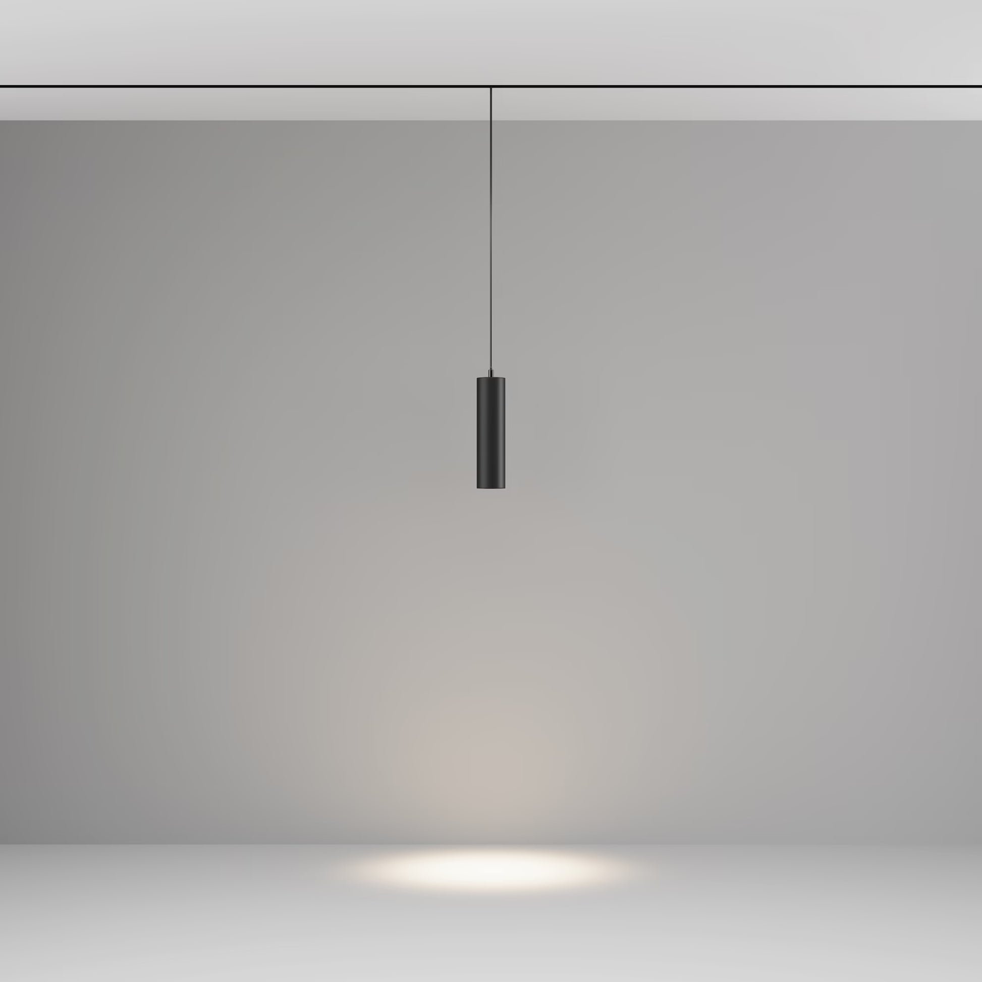 A modern black cylindrical track pendant light hanging from a thin cord against a minimalist gray studio background, casting a warm spotlight downward on the floor.