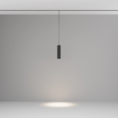 A modern black cylindrical track pendant light hanging from a thin cord against a minimalist gray studio background, casting a warm spotlight downward on the floor.