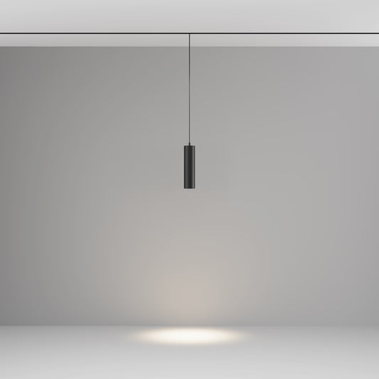 A modern black cylindrical track pendant light hanging from a thin cord against a minimalist gray studio background, casting a warm spotlight downward on the floor.