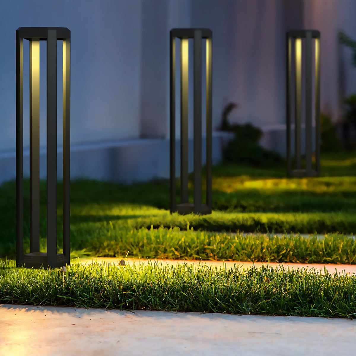 Three modern outdoor solar post lights with a dark metal frame are illuminated on a grassy pathway at dusk, casting a warm yellow glow.