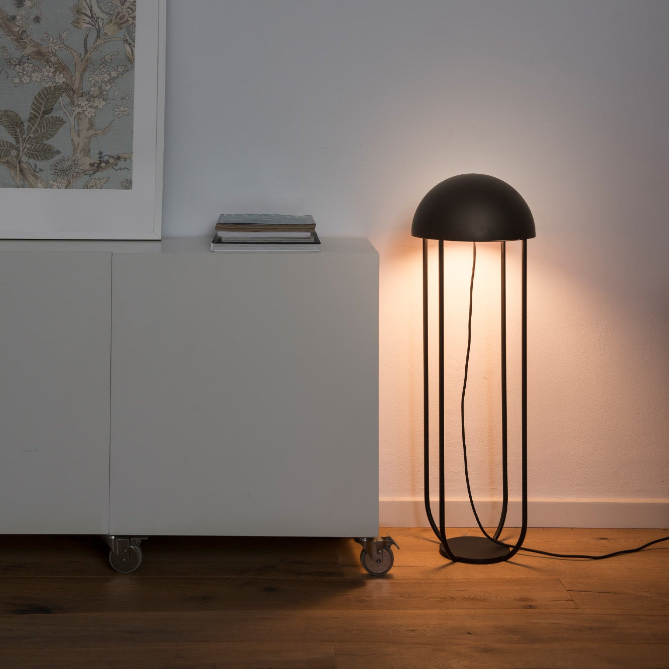 Modern black floor lamp next to a white cabinet with a framed picture on a plain wall.