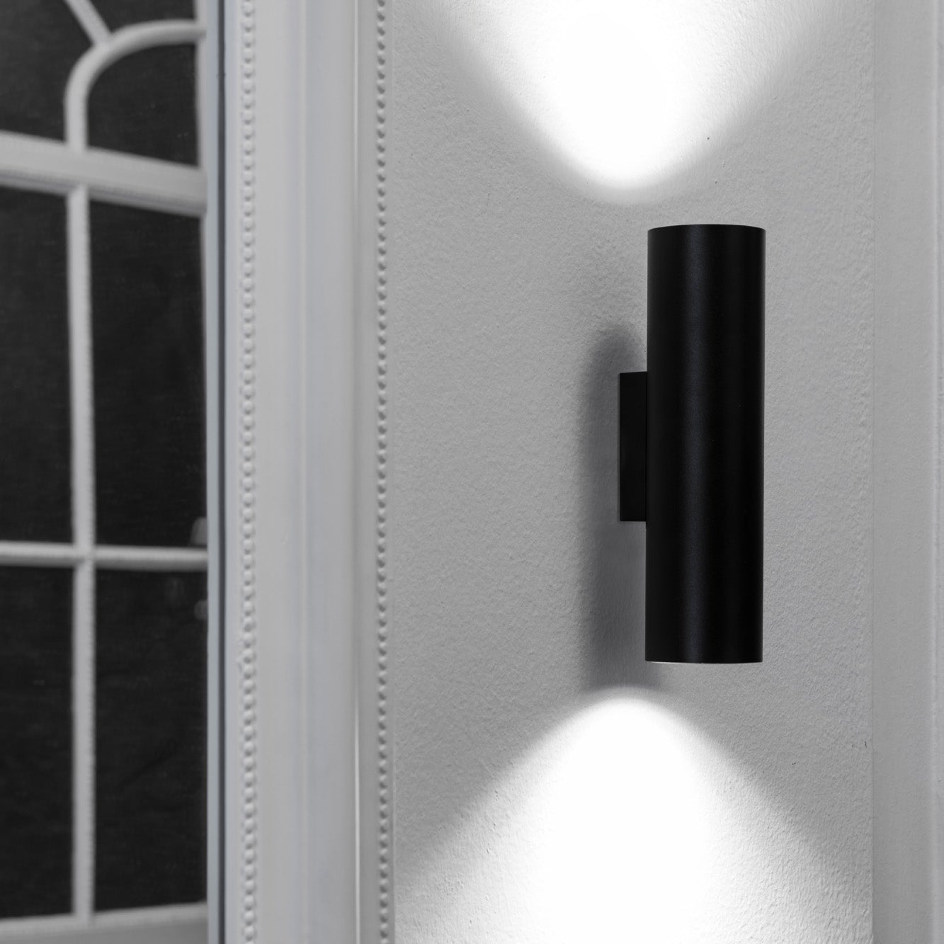 Black wall-mounted light fixture on a white wall with a window in the background