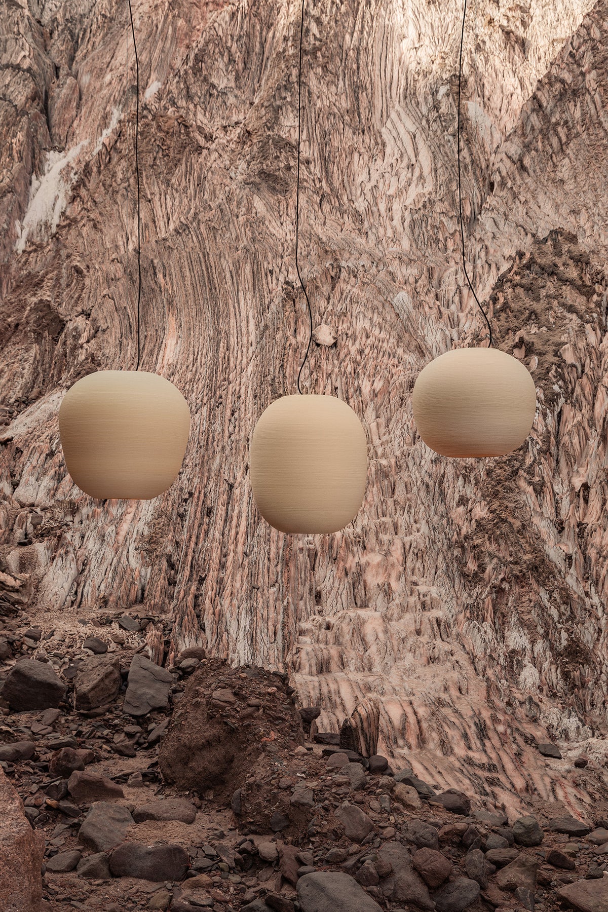 Three TIERRA 470 premium pendant lights suspended against a dramatic, textured rock cliffside. The earthy, rugged background perfectly highlights the organic, 3D-printed texture and warm beige tones of the sculptural shades.