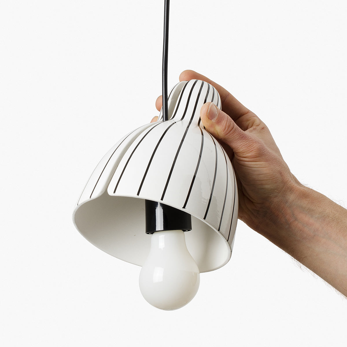 A hand holding a modern pendant light shade with a white and black striped pattern, featuring a black hanging cord and a white bulb.
