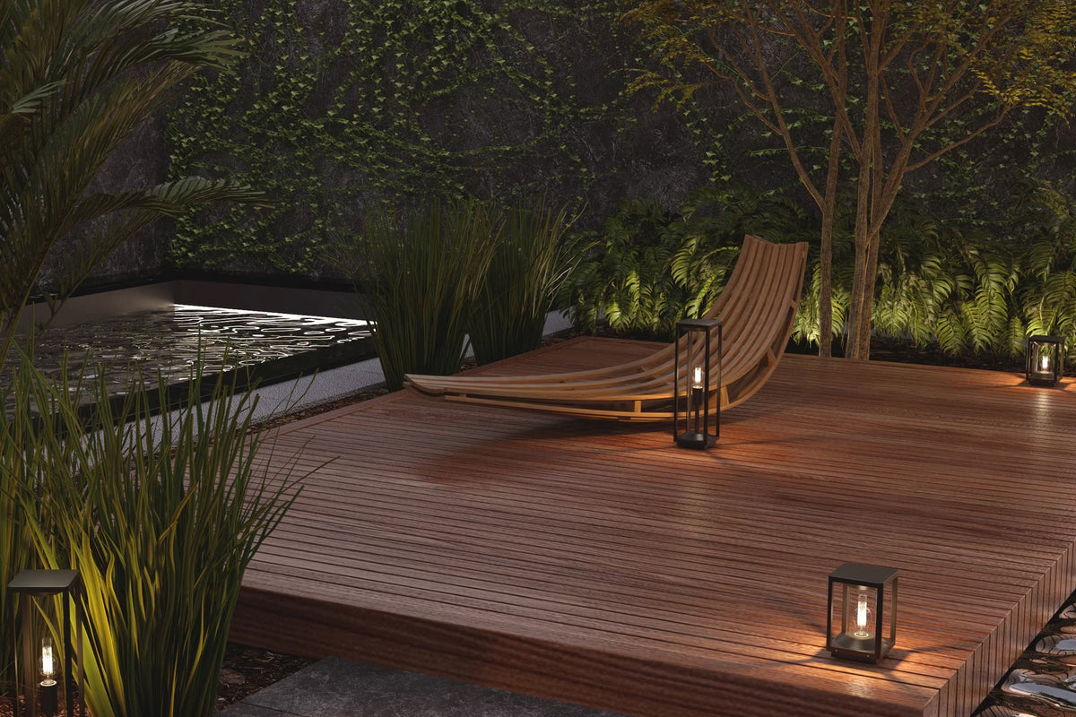 A modern wooden deck with a curved lounge chair, surrounded by lush green plants and illuminated by warm lanterns at night.